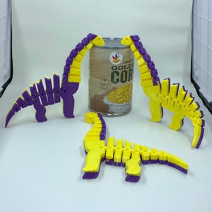 May include: Three yellow and purple dinosaur toys made of plastic are arranged around a can of corn. The can is labeled "Whole Kernel Golden Corn".