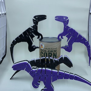 May include: Three articulated dinosaur figures in black and purple, with white accents. The dinosaurs are posed around a can of whole kernel corn. The dinosaurs are made of multiple pieces that fit together.