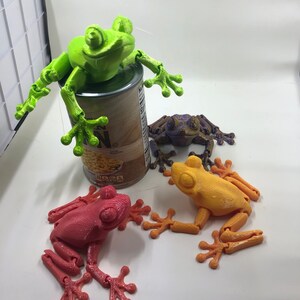 May include: Three articulated frog figurines, one green, one purple, and one orange, are posed on a white surface. The green frog is perched on top of a can of corn.
