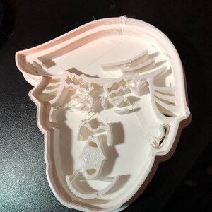 May include: A white plastic cookie cutter in the shape of a face with a distinctive hairstyle. The cutter has a layered design, with the facial features and hair defined by raised edges. The overall shape is a recognizable portrait.