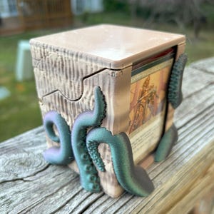 Op de afbeelding: Een beige, 3D-geprinte kaartenopbergdoos met een deksel, met een houtnerftextuur en teal en paarse tentakels. De doos is ontworpen om ruilkaarten in op te bergen, met enkele zichtbaar aan de binnenkant.