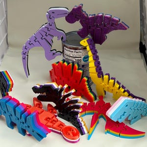 May include: A collection of colorful, articulated dinosaur puzzles in various colors, including purple, red, yellow, and blue. Some puzzles are assembled, while others are partially constructed. A can of food is in the background.