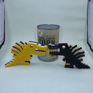 May include: Two 3D printed dinosaur figures, one yellow and one black, with purple and white accents. The dinosaurs are facing each other and are positioned in front of a can of whole kernel golden corn.