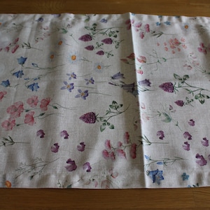 May include: A beige linen table runner with a floral pattern featuring pink, purple, and blue flowers. The runner is on a wooden table.