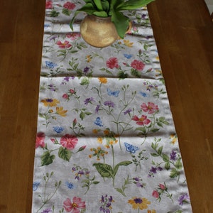 Peut inclure: Un chemin de table blanc avec un motif floral composé de fleurs roses, jaunes et bleues. Un bouquet de tulipes rouges dans un vase en céramique brun est placé sur le chemin de table.