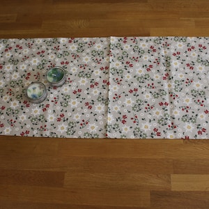 May include: A floral table runner with a light gray background, adorned with red, white, and green flower patterns. Two small, round candles sit on the runner. The runner is displayed on a wooden surface.