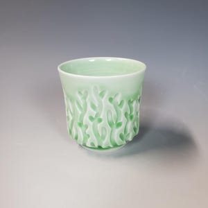 May include: A light green ceramic cup with a textured, raised pattern of organic shapes. The cup has a slightly flared rim and a small, circular base. The cup is approximately 3.5 inches tall and 3 inches in diameter.