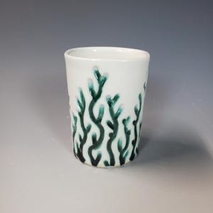 May include: White ceramic tumbler with a teal, coral-like design. The design is raised and wraps around the exterior of the cup. The cup has a smooth, glossy finish and a simple, cylindrical shape. The interior is plain white.