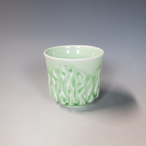 May include: A light green ceramic cup with a textured, organic pattern around the lower half. The cup has a smooth, glossy finish and a slightly flared rim. The design resembles stylized plant forms. The cup is approximately 3.5 inches tall.