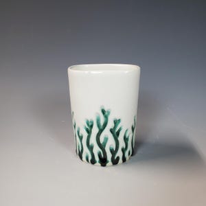 May include: A white ceramic tumbler with a dark teal, coral-like pattern around the base. The cylindrical cup has a smooth, glossy finish and a simple, modern design. The teal pattern adds a touch of color and visual interest.