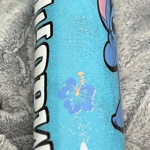 Op de afbeelding: Een lichtblauwe tumbler met een witte glitterafwerking. De tumbler heeft een cartoonfiguur met blauwe vacht en een roze oor, samen met twee roze en blauwe hibiscusbloemen. Het woord "Aloha" staat in wit op de zijkant van de tumbler.