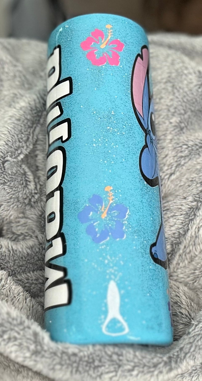 Op de afbeelding: Een lichtblauwe tumbler met een witte glitterafwerking. De tumbler heeft een cartoonfiguur met blauwe vacht en een roze oor, samen met twee roze en blauwe hibiscusbloemen. Het woord "Aloha" staat in wit op de zijkant van de tumbler.