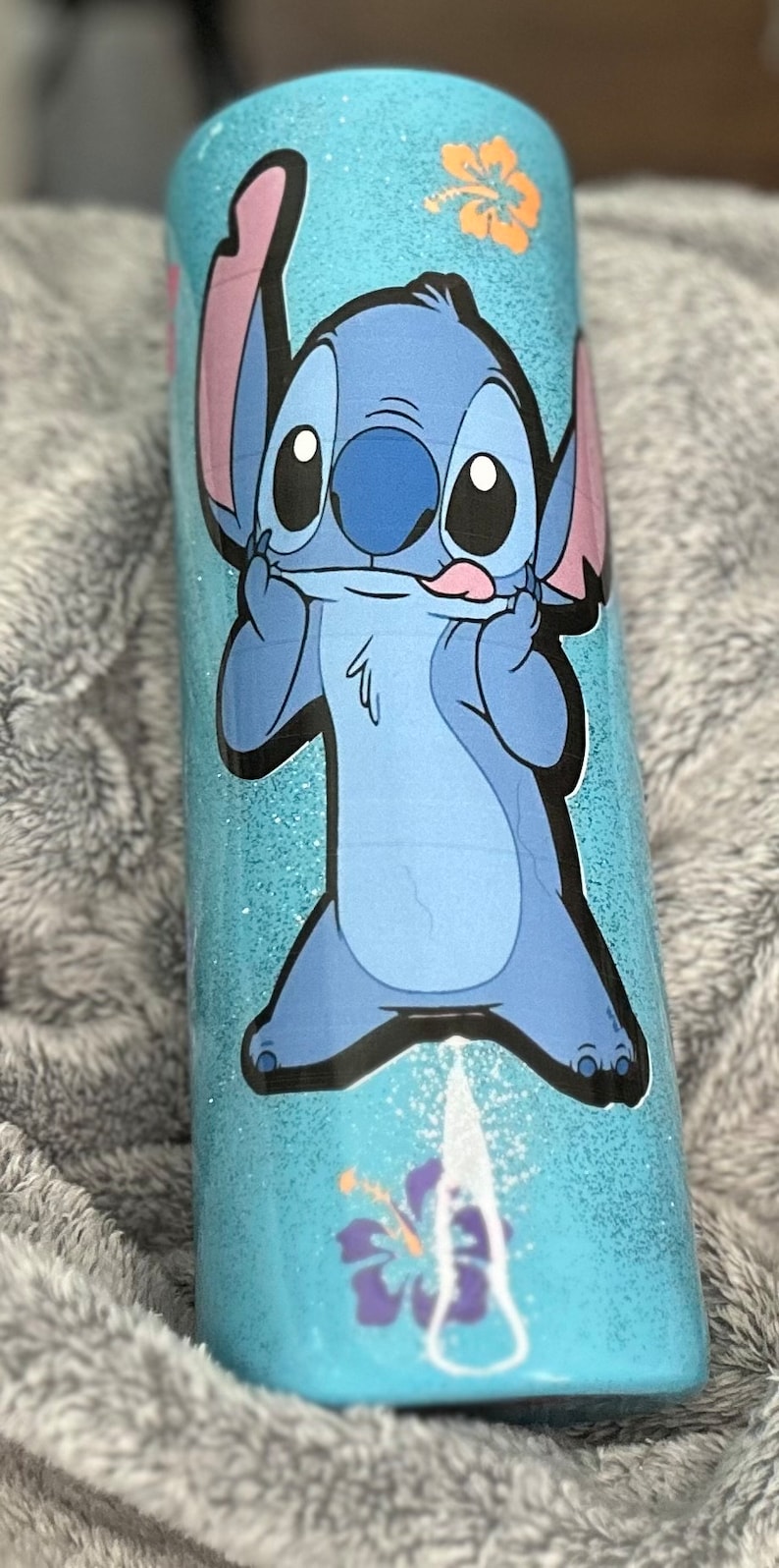 Op de afbeelding: Een blauwe en witte tumbler met een cartoon afbeelding van Stitch uit Lilo & Stitch. De tumbler heeft een glitter afwerking en een bloemenontwerp.