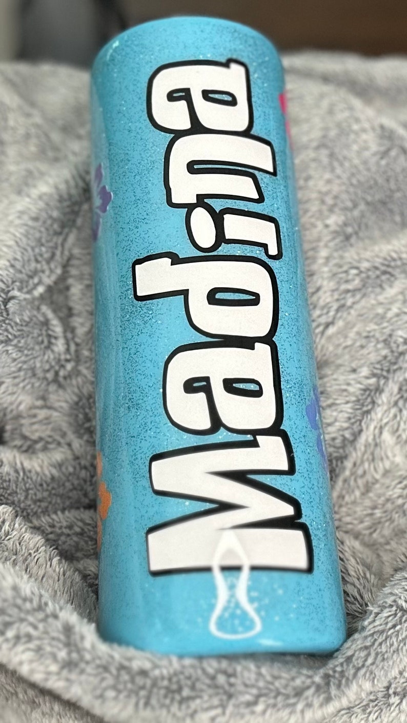 Op de afbeelding: Een lichtblauwe tumbler met een witte glitterafwerking en het woord "Mama" in wit met zwarte contouren. De tumbler heeft verschillende kleurrijke bloemen en potafdrukken rond het woord.