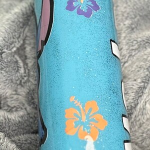 Op de afbeelding: Een lichtblauwe tumbler met een glitterafwerking. De tumbler heeft een cartoonfiguur en twee oranje en paarse hibiscusbloemen.