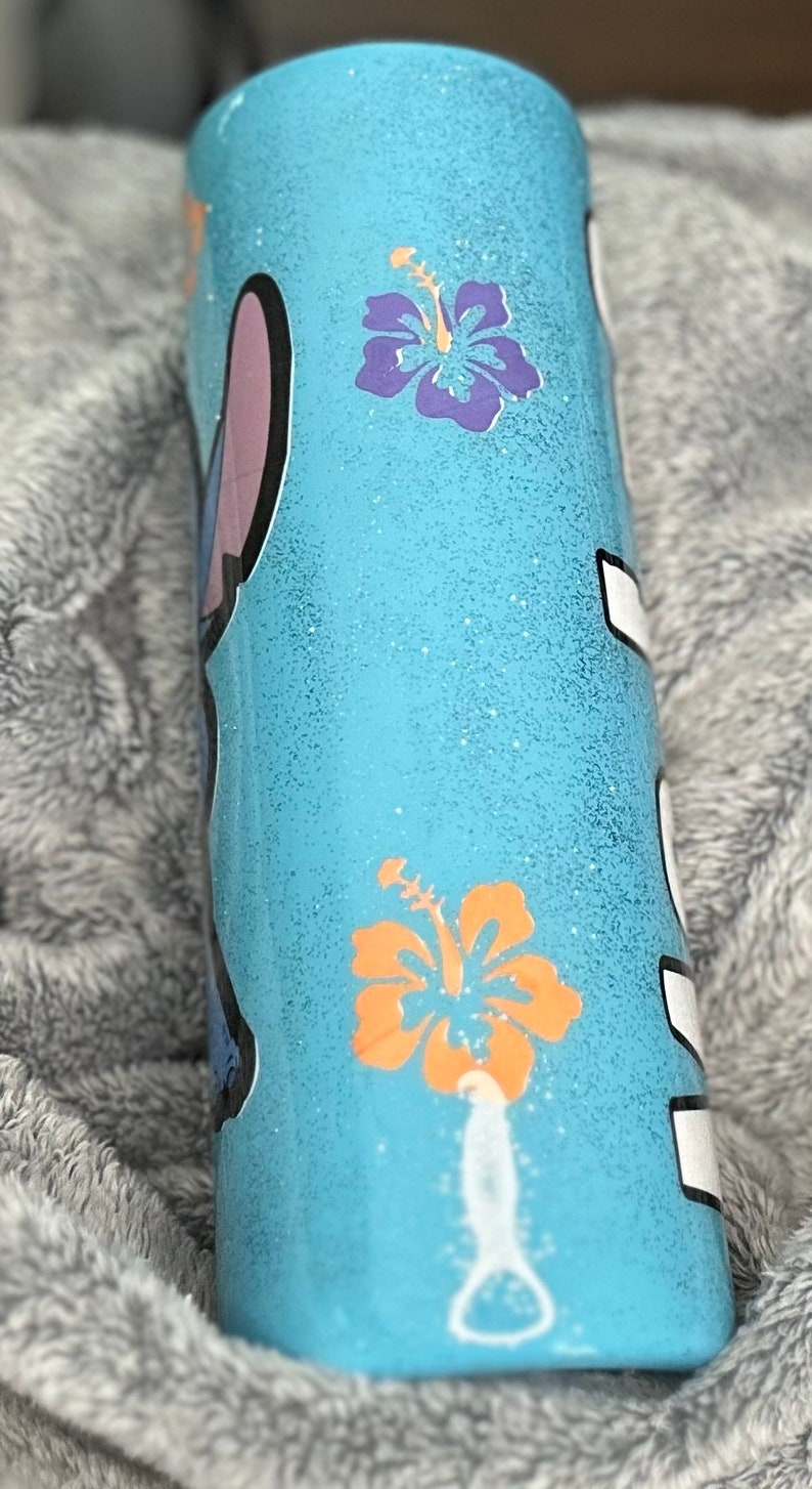 Op de afbeelding: Een lichtblauwe tumbler met een glitterafwerking. De tumbler heeft een cartoonfiguur en twee oranje en paarse hibiscusbloemen.