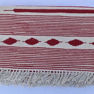 May include: A rectangular, woven rug with a cream-colored background and horizontal red stripes. The rug features a central band of red diamond shapes and a fringe along the bottom edge. The rug measures approximately 60 inches long.
