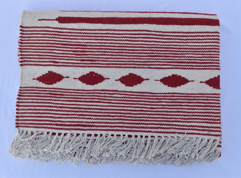 May include: A rectangular, woven rug with a cream-colored background and horizontal red stripes. The rug features a central band of red diamond shapes and a fringe along the bottom edge. The rug measures approximately 60 inches long.