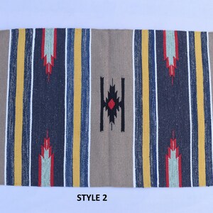 Puede incluir: Una alfombra rayada con un patr&oacute;n geom&eacute;trico en el centro. La alfombra tiene un fondo beige con rayas amarillas, azules y rojas. El patr&oacute;n geom&eacute;trico es negro con un diamante rojo en el centro. La alfombra tiene flecos blancos en los bordes. El texto "STYLE 2" est&aacute; impreso debajo de la alfombra.