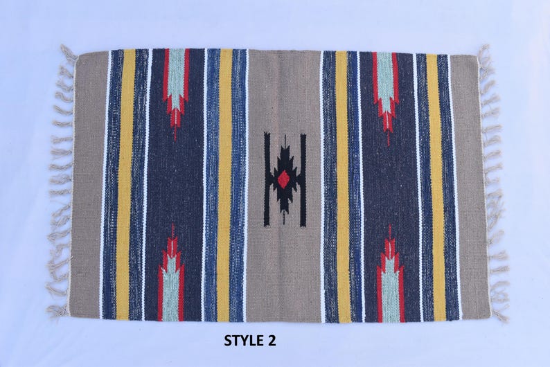Puede incluir: Una alfombra rayada con un patr&oacute;n geom&eacute;trico en el centro. La alfombra tiene un fondo beige con rayas amarillas, azules y rojas. El patr&oacute;n geom&eacute;trico es negro con un diamante rojo en el centro. La alfombra tiene flecos blancos en los bordes. El texto "STYLE 2" est&aacute; impreso debajo de la alfombra.