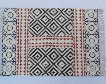 May include: A rectangular cotton rug with a geometric pattern in black, white, and red. The rug has a fringe border and is made of natural fibers.
