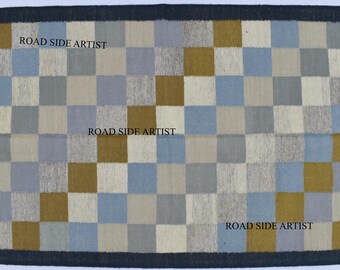 May include: A rectangular rug with a checkered pattern in shades of blue, gray, beige, and gold. The rug has a dark blue border and fringe along the edges. The text "ROAD SIDE ARTIST" is printed on the rug.