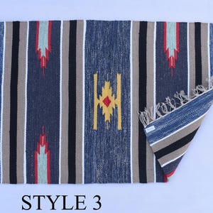 Puede incluir: Una alfombra rayada azul, beige, negro y rojo con un patr&oacute;n geom&eacute;trico. La alfombra tiene flecos en todos los lados. "STYLE 3" est&aacute; escrito debajo de la alfombra.
