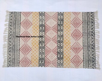 May include: A rectangular rug with a geometric pattern in shades of red, yellow, and black. The rug has a white background and is made of cotton. The rug has fringe on all four sides.