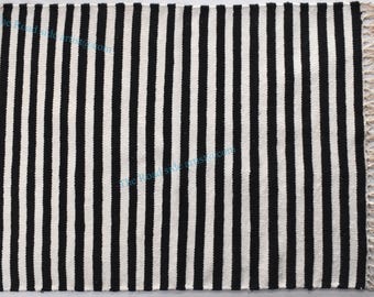 May include: Black and white striped rug with fringe on all sides. The rug is made of a woven material and has a simple, modern design.