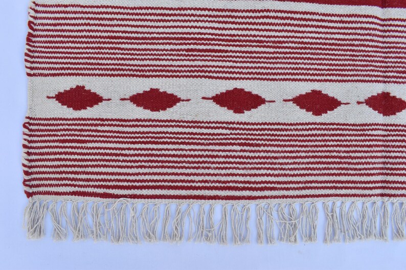 May include: A woven rug with a red and white striped pattern. The rug features a series of red diamond shapes along the center and a fringe along the bottom edge. The rug is made of a textured material.