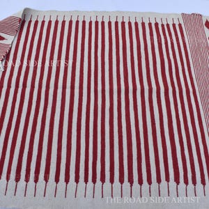 May include: A rectangular rug with a cream-colored background and vertical red stripes. The rug has a fringed edge and diamond-shaped patterns on the sides. The text "THE ROAD SIDE ARTIST" is visible.