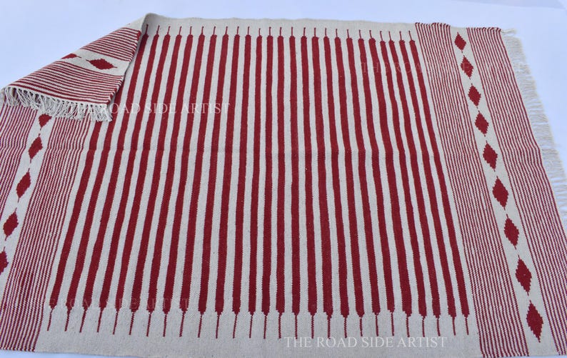 May include: A rectangular rug with a cream-colored background and vertical red stripes. The rug has a fringed edge and diamond-shaped patterns on the sides. The text "THE ROAD SIDE ARTIST" is visible.