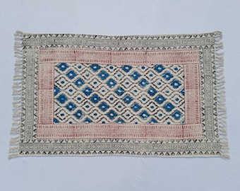 May include: A white cotton rug with a blue and black geometric pattern. The rug has a red border and fringe on all sides.
