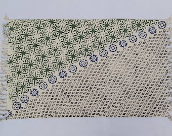 May include: A white cotton rug with a green and blue geometric pattern. The rug has a fringe border.