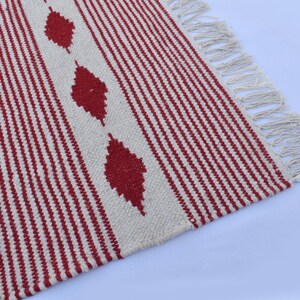 May include: A woven rug with a cream-colored background and vertical red stripes. The rug features three red diamond shapes and cream-colored fringe along one edge. The rug is made of a textured material.