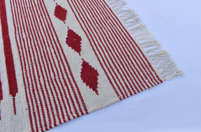 May include: A woven rug with a cream-colored background and vertical red stripes. The rug features three red diamond shapes and cream-colored fringe along one edge. The rug is made of a textured material.