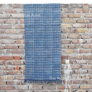 Peut inclure: Un textile bleu et blanc à motifs avec une bordure à franges est suspendu à un mur de briques. Le textile présente un motif de losanges répétitif. Le texte "The Road Side Artist" est visible.