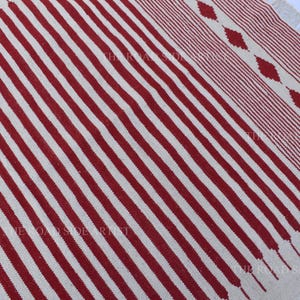 May include: A rectangular rug with a cream-colored background and bold, vertical red stripes. The rug's design includes a patterned border with diamond shapes and fringed edges, offering a textured look.