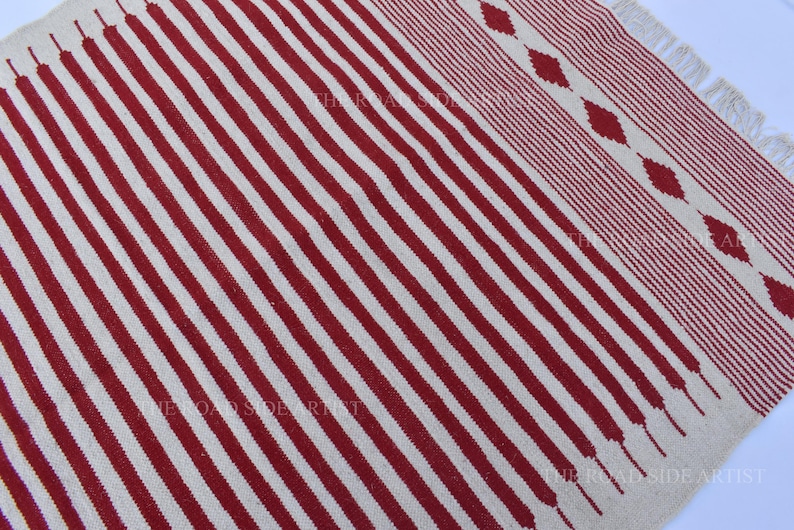 May include: A rectangular rug with a cream-colored background and bold, vertical red stripes. The rug's design includes a patterned border with diamond shapes and fringed edges, offering a textured look.