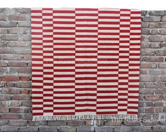 Puede incluir: Una alfombra a rayas rojas y blancas con un patrón geométrico cuelga de una pared de ladrillo. La alfombra tiene flecos en la parte inferior. El diseño presenta rayas horizontales rojas y blancas alternas.