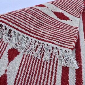 May include: A red and white striped rug with a fringed edge. The rug features a repeating pattern of red stripes and leaf-shaped designs on a white background. The rug is made of a woven material.