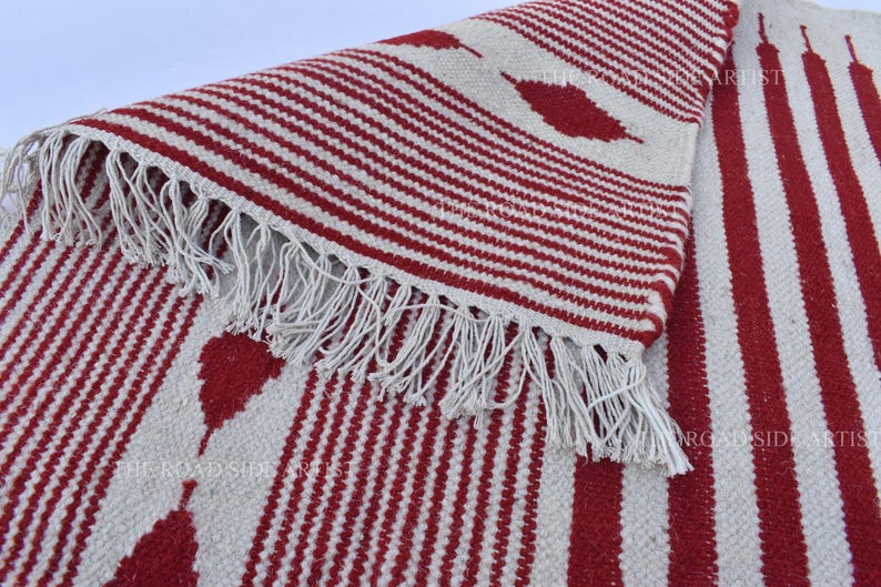 May include: A red and white striped rug with a fringed edge. The rug features a repeating pattern of red stripes and leaf-shaped designs on a white background. The rug is made of a woven material.