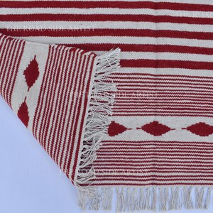 May include: A red and white striped rug with a geometric diamond pattern. The rug has a fringed edge and is partially rolled up, revealing the underside. The design features alternating stripes and diamond shapes.