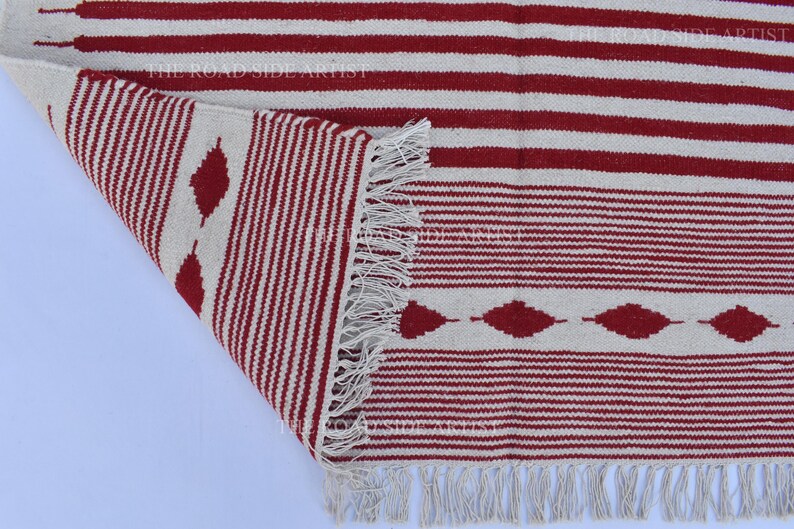 May include: A red and white striped rug with a geometric diamond pattern. The rug has a fringed edge and is partially rolled up, revealing the underside. The design features alternating stripes and diamond shapes.