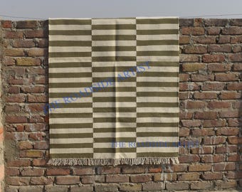 May include: A striped rug with alternating bands of white and olive green. The rug has a fringe border and is hanging on a brick wall.