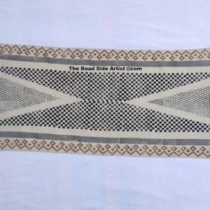 Puede incluir: Una alfombra con estampado geométrico en blanco y negro con bordes marrones y grises. La alfombra tiene flecos en todos los lados. El texto "The Road Side Artist @com" está impreso en la alfombra.