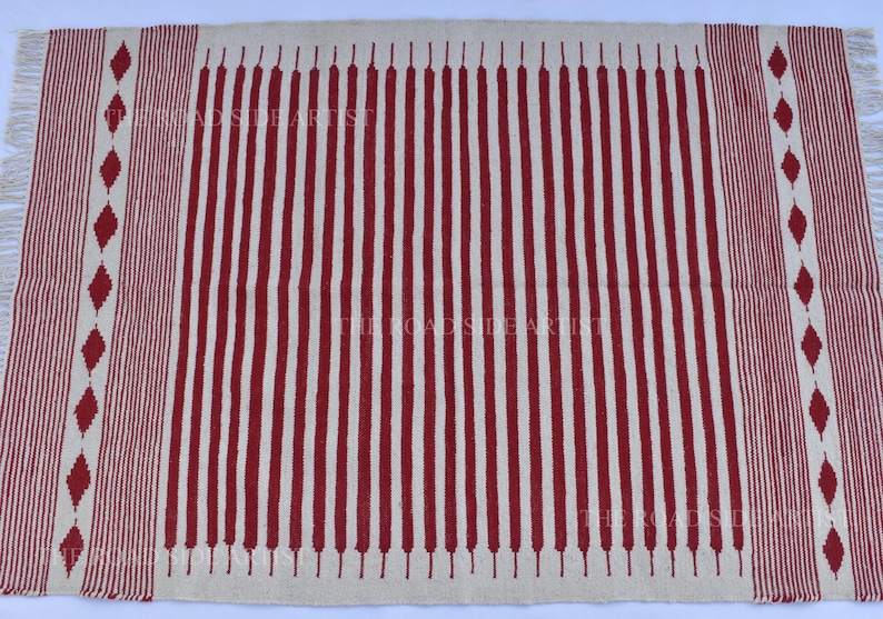 May include: A rectangular rug with a cream-colored background and vertical red stripes. The rug has a fringed edge and a decorative border with red diamond shapes. The text "THE ROADSIDE ARTIST" is visible.