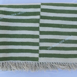 May include: Green and white striped rug with fringe. The rug has a woven texture and is made of natural fibers. The text "THE ROADSIDE ARTIST" is printed on the rug.