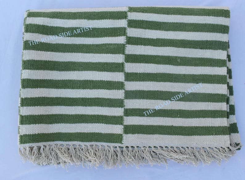 May include: Green and white striped rug with fringe. The rug has a woven texture and is made of natural fibers. The text "THE ROADSIDE ARTIST" is printed on the rug.
