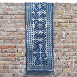 Puede incluir: Textil rectangular azul índigo con un estampado floral blanco y flecos. El diseño floral se repite en el centro, con un borde de puntos conectados. El textil se muestra contra una pared de ladrillos. Mide unos 150 cm de largo.